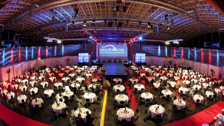 Large event room with round tables and stage, illuminated in red and blue.