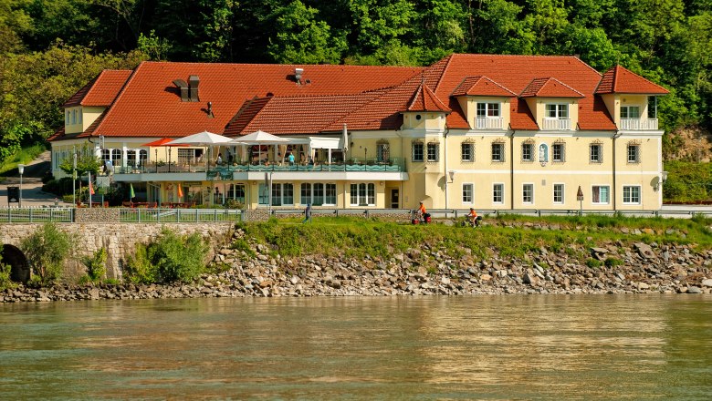 A yellow building with red roofs on the riverbank, surrounded by trees.