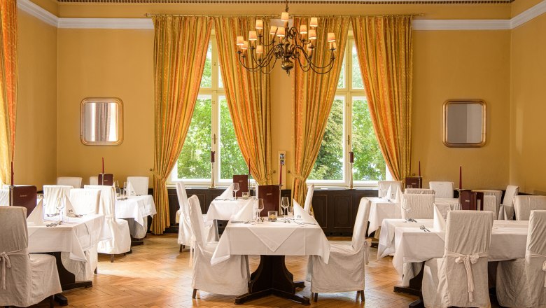 Elegant hotel restaurant with laid tables, white chair covers and large windows with yellow curtains.