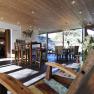 A modern breakfast room with wooden furniture, large windows and a chandelier. A wooden chair in the foreground and a dining table in the background.
