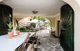 A covered passageway with a wooden table and plants leads to a paved courtyard with tables and chairs.