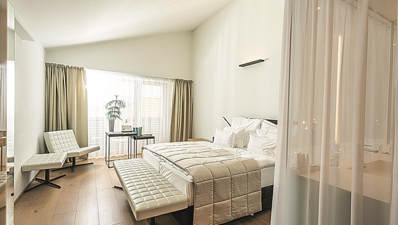 Modern hotel room with large bed, white furniture and wooden floor.