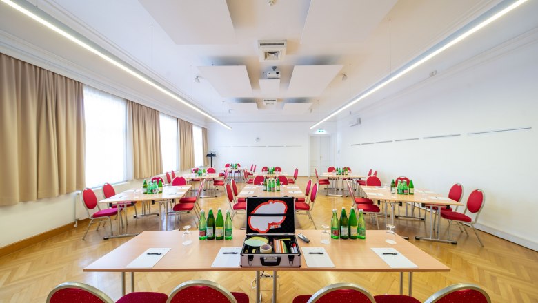 A bright seminar room with red chairs and tables on which water bottles and writing materials are placed.