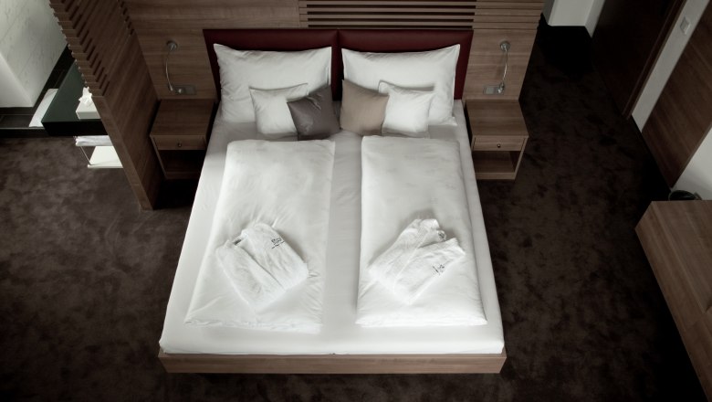 A modern hotel room with a double bed, white comforters and bathrobes, surrounded by wooden furniture.