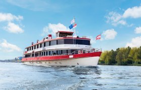A passenger ship with red and white colors sails on a river under a blue sky.