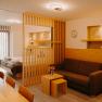A modern hotel apartment with a double bed, light-colored wood furniture, a pull-out couch, and a wooden room divider. Part of the wall is paneled with wood, and there are niches in the wall painted in an earthy gold.