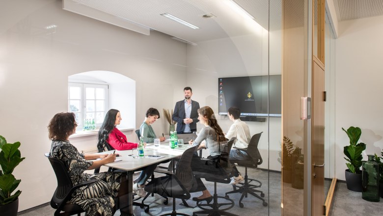 A modern conference room with six people sitting at a table listening to a speaker. A screen in the background.