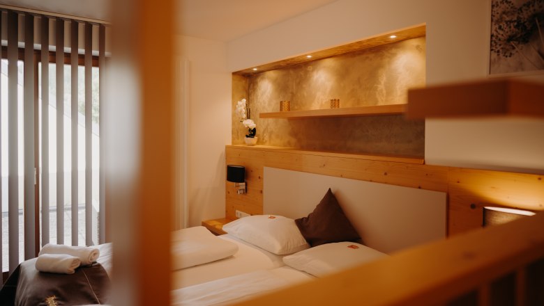 A cozy apartment with a double bed and a balcony door. Part of the wall is paneled with light-colored wood; behind the bed is a recessed niche with a shelf and spotlights, painted in an earthy gold.