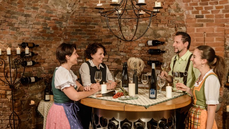 Four people in traditional dress are standing in a wine cellar holding wine glasses. They are laughing and chatting at a round table.