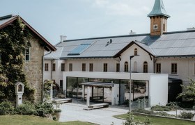 A modern building with solar panels on the roof and a clock tower, surrounded by green spaces.