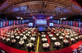 Large event room with round tables and stage, illuminated in red and blue.