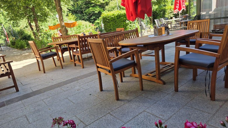 A cozy outdoor dining area with wooden furniture and red umbrellas, surrounded by green trees and flowers.