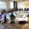 People sit in a circle on cushions and chairs in a bright seminar room with a wooden floor.