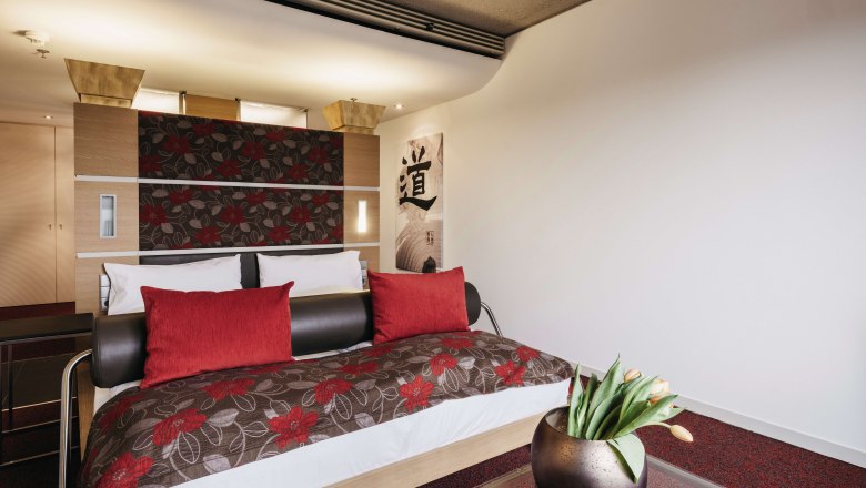 Modern hotel room with double bed, red cushions and floral pattern. A glass coffee table with a vase full of tulips stands in the foreground.