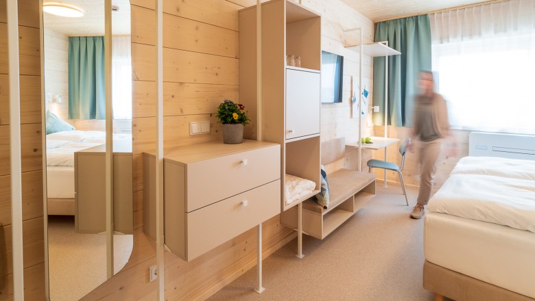 Modern hotel room with wooden paneling, mirror, wardrobes and a bed. One person moves around the room.