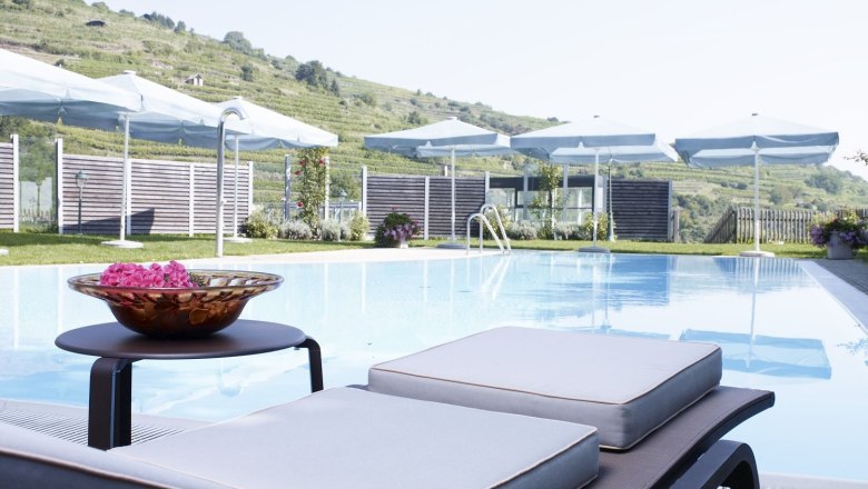 An outdoor pool with sun loungers and parasols in front of a hilly landscape.