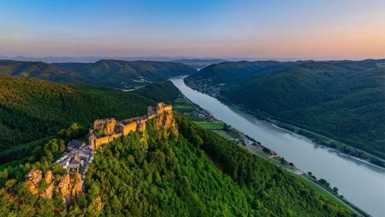 Aerial view of the ruins of Aggstein Castle on the Danube, surrounded by green hills and forests at sunset.