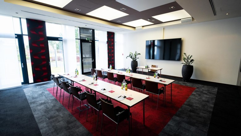 A modern conference room with tables, chairs, and a large screen on the wall. The room has large windows and plants as decoration.