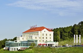 Landzeit Steinhäusl building with red roof and glass facade, surrounded by trees and blue sky.
