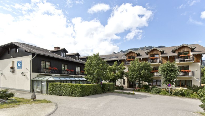 A traditional inn with wooden balconies and flower boxes, surrounded by trees and mountains in the background.