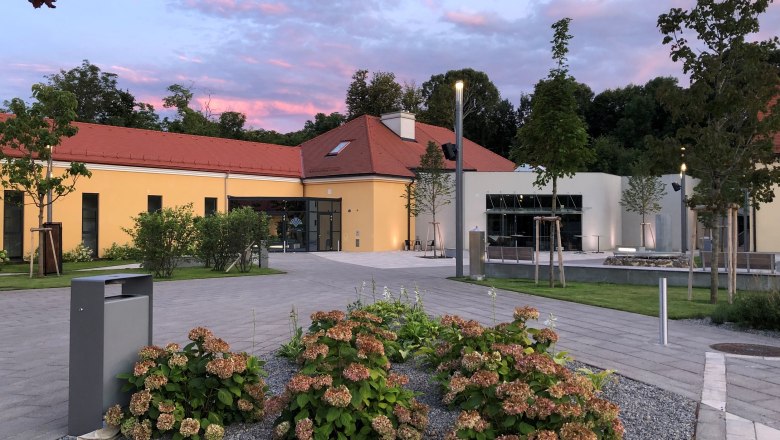 Modern building with yellow façade and red roof, surrounded by plants and trees, at sunset.