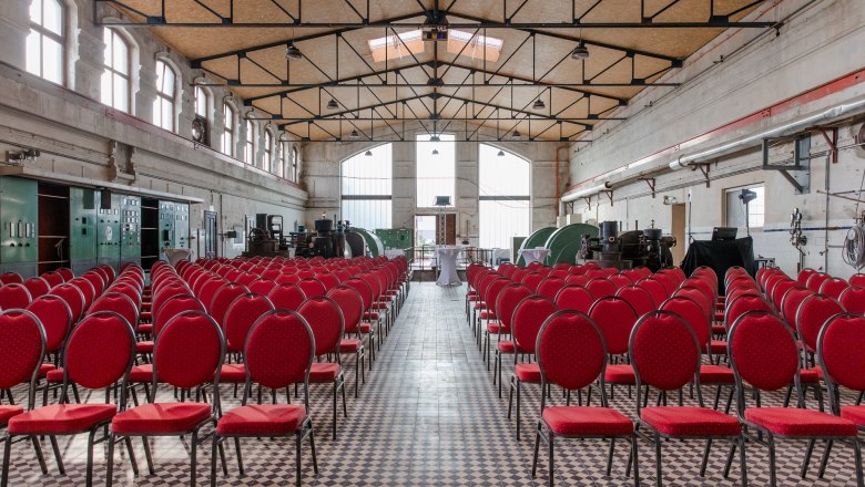 Interior view of an old turbine hall with red chairs and industrial machines.