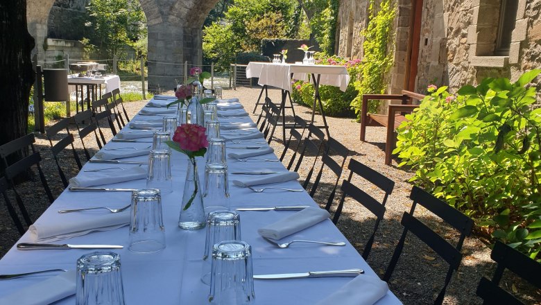 Long outdoor table with white tablecloth, set with glasses and cutlery, stone arches and green vegetation in the background.