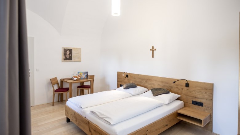 A simply furnished guest room with a double bed, wooden furniture and a cross on the wall.