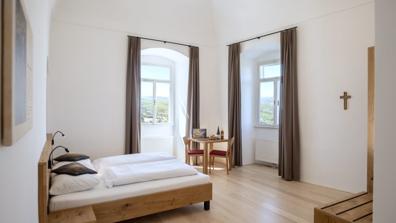 Bright double room with wooden bed, table and chairs, two windows with curtains and a cross on the wall.