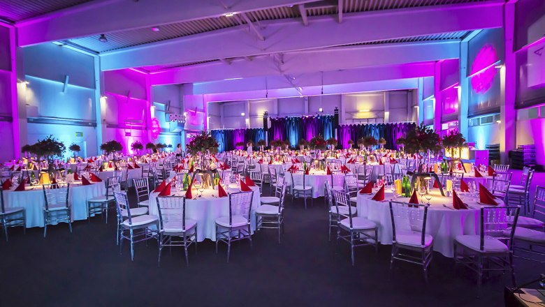 Festively decorated hall with round tables, white tablecloths and purple lighting.