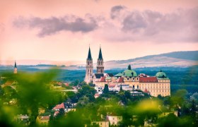 Panoramic view of Stift Klosterneuburg with two church towers and baroque building, surrounded by green landscape and under a cloudy sky.