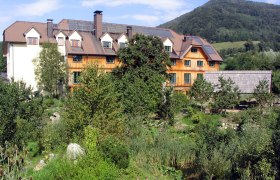 A large building with a wooden fa&ccedil;ade, surrounded by lush greenery and a mountain in the background.