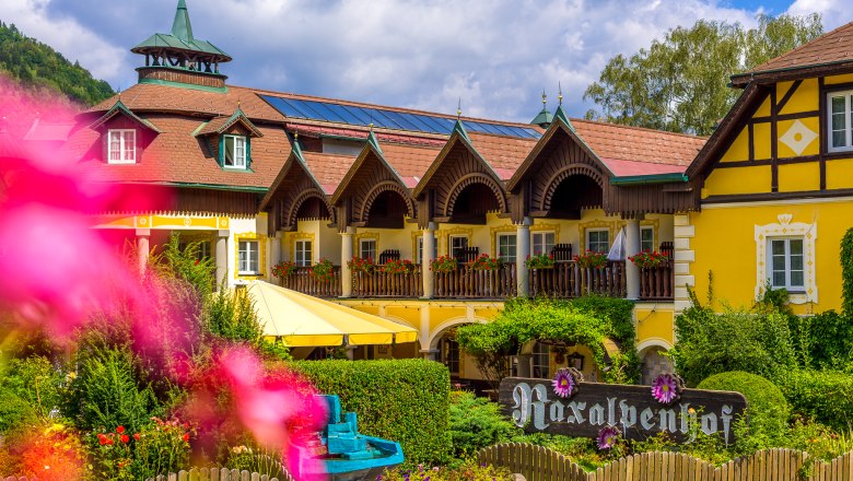 Raxalpenhof Hotel, © Wiener Alpen / Christian Kremsl A yellow, traditional hotel with wooden balconies and flowers, surrounded by green trees and a blue sky.