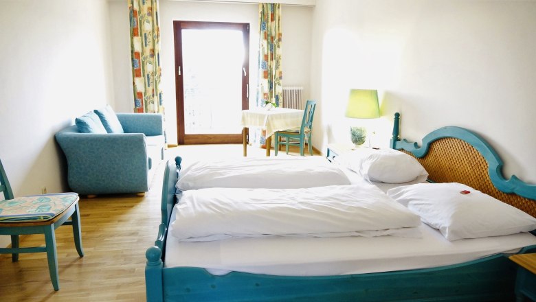 A bright room with a double bed, blue couch, table and chair in front of a window with curtains.