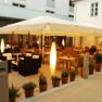 An illuminated guest garden with tables, chairs and plants in pots, surrounded by buildings.