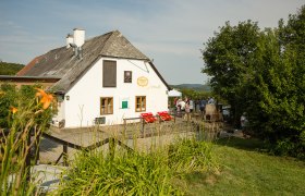 White country house with shingle roof, surrounded by green landscape and people in the background.