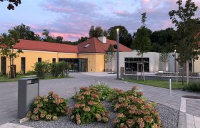 Modern building with yellow façade and red roof, surrounded by plants and trees, at sunset.