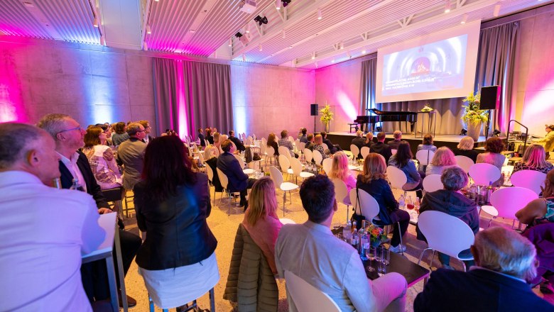 People sit in a modern event room with a stage and screen.