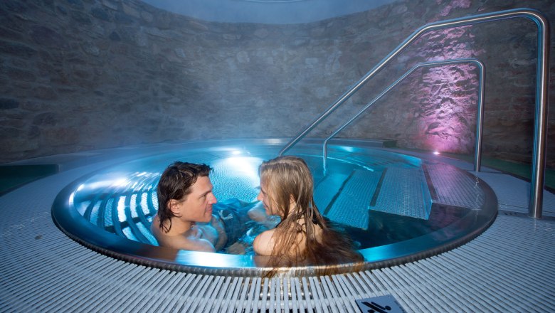 A couple relaxes in a round whirlpool with blue lighting and stone walls.