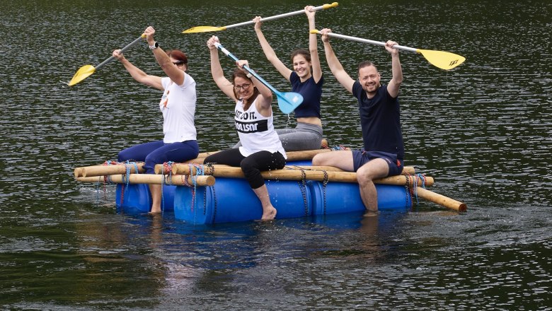 Four people sit on a homemade raft made of blue barrels and wooden poles on a lake and happily lift their paddles into the air.