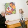Two priests talk to two children in a gallery in front of a colorful painting.