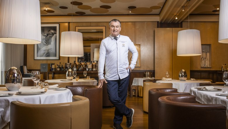 A man in a white chef's jacket stands in an elegant restaurant with round tables and large lamps.