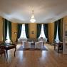 Elegant room with wooden floor, green curtains, two armchairs, a sofa and a desk. A chandelier hangs from the ceiling.