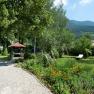 A well-kept garden with a gravel path, flower beds, a pavilion and a lounger. Trees and mountains can be seen in the background.