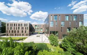 Modern university buildings with a brown façade and large windows, surrounded by green vegetation under a blue sky.