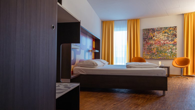 Modern hotel room with bed, yellow curtains and colorful painting on the wall.