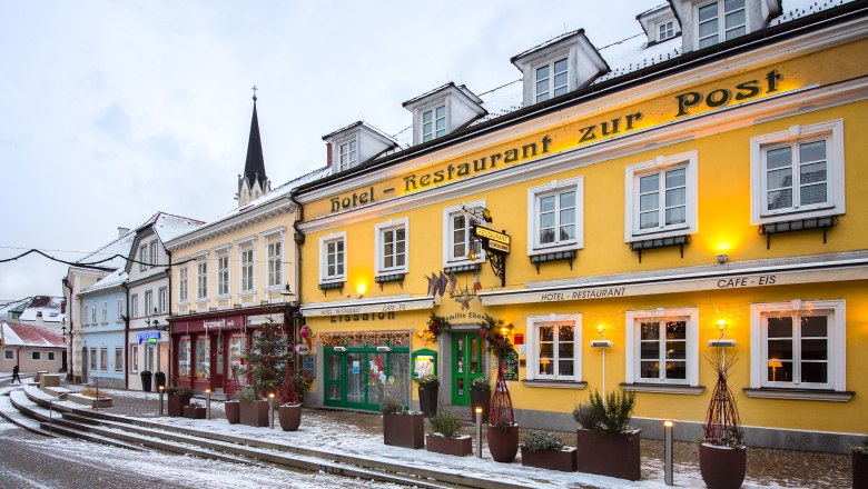 Exterior view of a yellow building with the inscription 'Hotel-Restaurant zur Post' in winter, surrounded by snow and decorated with lights.