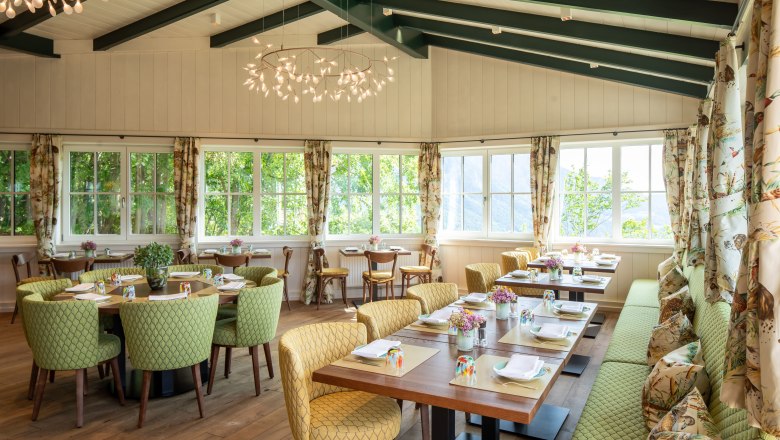 A bright conservatory with covered tables, green chairs and large windows.