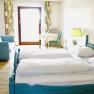 A bright room with a double bed, blue couch, table and chair in front of a window with curtains.