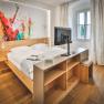 A modern hotel room with a double bed, an abstract artwork on the wall, and a TV on a shelf.